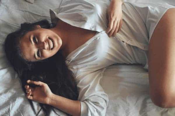 Femme détendue et souriante allongée sur un lit, symbole de plaisir, de confiance et de reconnexion au corps féminin.